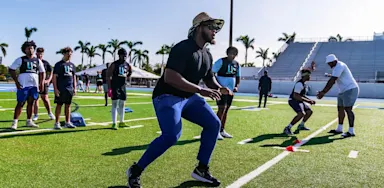 A football coach in a sun hat leads student-athletes through cone drills on a bright green turf field, while others watch and wait their turn during outdoor training at IMG Academy.