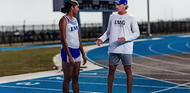 An IMG Academy coach speaks with a student-athlete on a track, giving instruction while they stand side by side near the starting lanes.