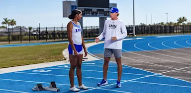 A coach speaks with an athlete beside starting blocks on a track.