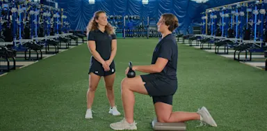 An athlete kneels in a lunge with a kettlebell beside a trainer.