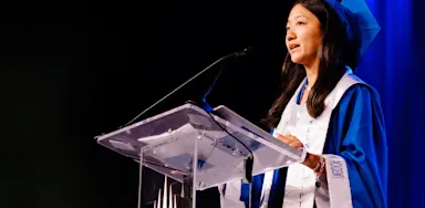 Young female graduate delivering a commencement speech in a bright blue cap and gown at a graduation ceremony podium.