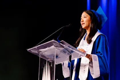Young female graduate delivering a commencement speech in a bright blue cap and gown at a graduation ceremony podium.