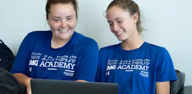 Two IMG Academy female student-athletes sit together on a couch, smiling at a laptop screen while working on a digital project or reviewing materials online.
