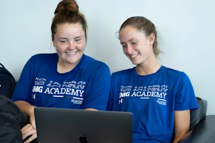 Two IMG Academy female student-athletes sit together on a couch, smiling at a laptop screen while working on a digital project or reviewing materials online.