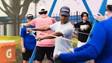An IMG Academy golfer performs a resistance band drill, pulling a band across his body while focusing on form, with other athletes training in the background near a practice area.