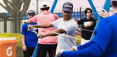 An IMG Academy golfer performs a resistance band drill, pulling a band across his body while focusing on form, with other athletes training in the background near a practice area.