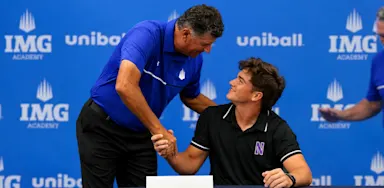 A seated student-athlete shakes hands with an adult at a signing table in front of an IMG Academy backdrop.
