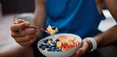 A close view shows an athlete eating a bowl of yogurt, fruit, and grains.