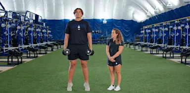 An athlete holds dumbbells at his sides while a trainer observes on indoor turf.