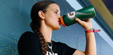 IMG Academy athlete drinks from a water bottle while seated indoors, wearing an IMG Academy shirt and braid hairstyle, taking a hydration break during training.