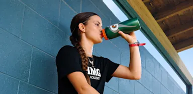 An athlete drinks from a green bottle beside an indoor court.