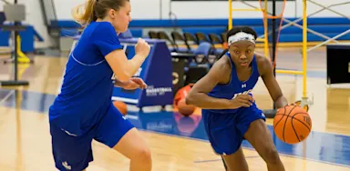 Two female basketball players practice on an indoor court, with one dribbling the ball while driving past a defender during a drill.
