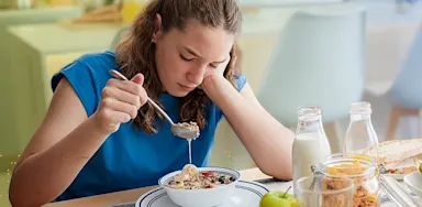 Teen athlete sits at a breakfast table looking hesitant while eating a bowl of cereal, illustrating nutrition challenges addressed in the “Performance Fuel for Picky Eaters” workshop.