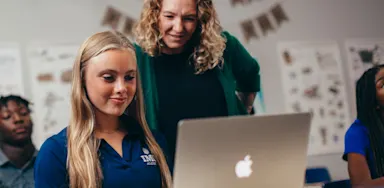 Student-athlete researching colleges on a laptop with guidance from a teacher, illustrating the support available during the college search process.