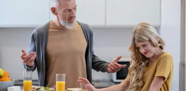 Frustrated father with his young, long-haired daughter who is pushing away a healthy meal (sandwich) in a modern kitchen, illustrating picky eating challenge.