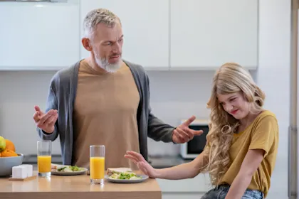 Frustrated father with his young, long-haired daughter who is pushing away a healthy meal (sandwich) in a modern kitchen, illustrating picky eating challenge.