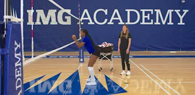 A volleyball player practices blocking at the net while a coach observes.