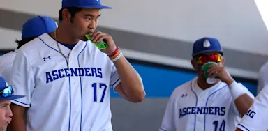 IMG Academy baseball players wearing white uniforms and blue caps hydrate and fuel with drinks and snacks in the dugout during a break in competition or practice.
