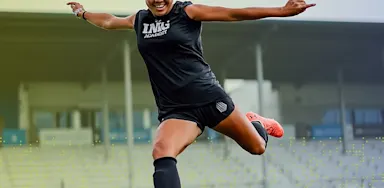 An IMG Academy student-athlete soccer player leaps into the air to strike the ball, extending her leg in a dynamic kicking motion on the field.