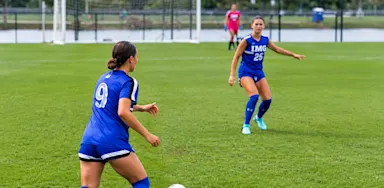 IMG Academy female soccer players in blue jerseys on a grass field, focused on a game play.