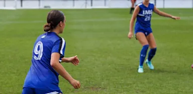 IMG Academy female soccer players in blue jerseys on a grass field, focused on a game play.