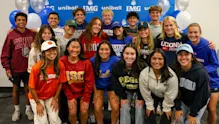 Group of IMG Academy student-athletes posing in college gear during a signing day event, celebrating commitments to various NCAA universities.