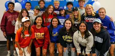 Group of IMG Academy student-athletes posing in college gear during a signing day event, celebrating commitments to various NCAA universities.