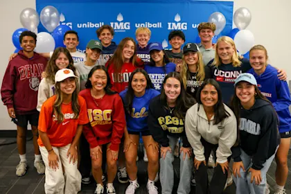 Group of IMG Academy student-athletes posing in college gear during a signing day event, celebrating commitments to various NCAA universities.
