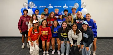A group of student-athletes poses in college shirts in front of an IMG Academy backdrop.