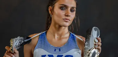 A close-up portrait of a serious-looking teenage girl wearing a white and blue IMG Academy track uniform. She has long brown hair tied back and is holding a pair of running spikes over her shoulders, with the gold cleats facing forward. She looks confidently into the camera against a dark studio background.