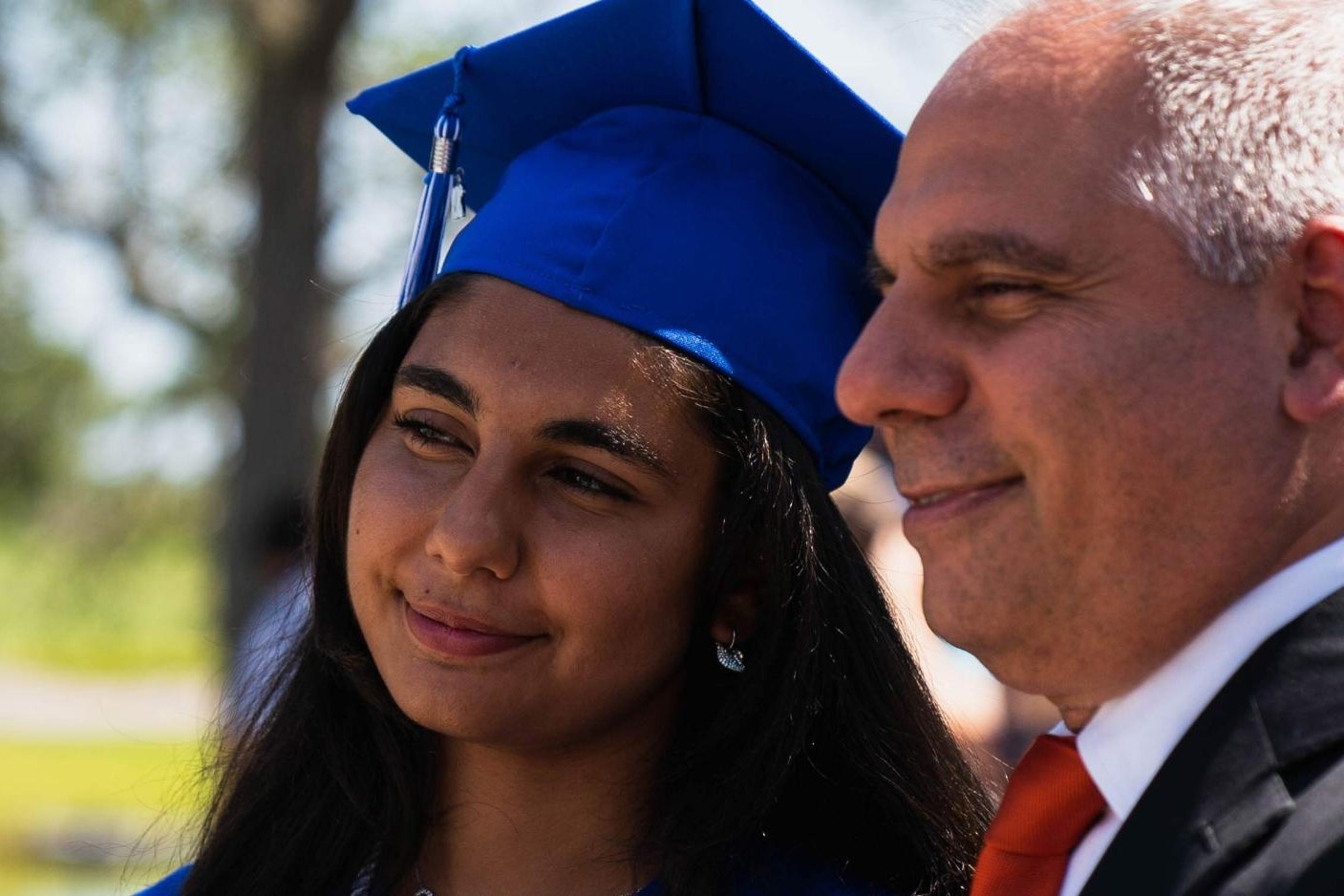 A young woman in a blue graduation cap and gown smiles while standing next to father in a suit, outdoors on a sunny day.