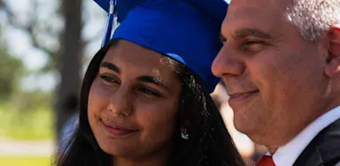 A young woman in a blue graduation cap and gown smiles while standing next to father in a suit, outdoors on a sunny day.