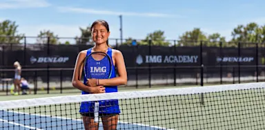 A tennis player stands smiling behind the net on an outdoor court.