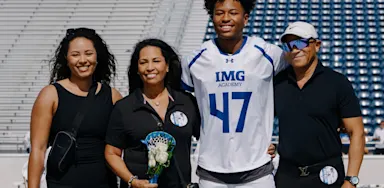 Lacrosse athlete (jersey #47) smiling with his family (parents and sister) on the turf during Senior Night ceremonies at IMG Academy Field.