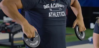 A close view shows an athlete holding weight plates during a shoulder exercise.
