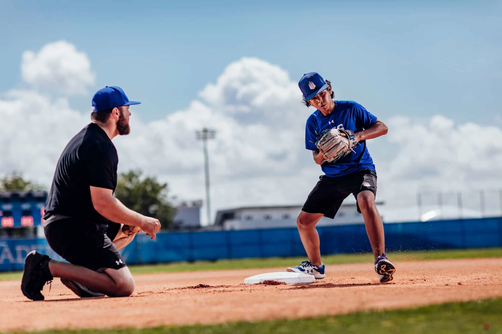 A baseball coach kneels on the infield during a sunny day as a young athlete wearing a blue shirt and cap practices fielding at second base, kicking up dirt mid-play with intense focus.