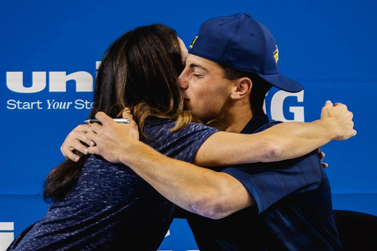 A young male athlete hugs a woman during a signing event, with a blue IMG Academy-branded backdrop behind them.