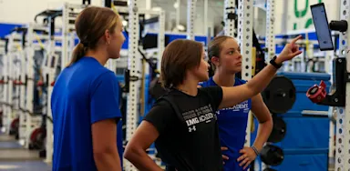 Three IMG Academy student-athletes stand in the weight room, looking at a digital screen mounted on a training rack to review strength metrics, workout plans, and overall goals.