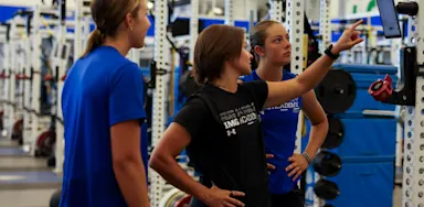 Three IMG Academy student-athletes stand in the weight room, looking at a digital screen mounted on a training rack to review strength metrics, workout plans, and overall goals.