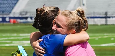 A mom and female athlete sharing an emotional hug on a sports field during a graduation or senior night ceremony.