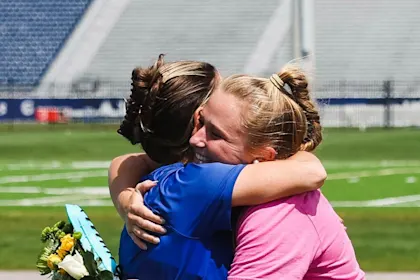 A mom and female athlete sharing an emotional hug on a sports field during a graduation or senior night ceremony.