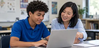 A student and a teacher look at a laptop together in a classroom.