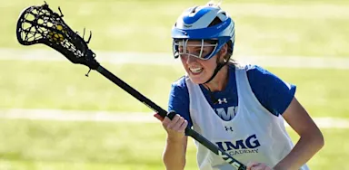 Female lacrosse player from IMG Academy cradling the ball during a game on a sunny field.