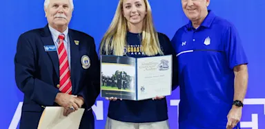 A female student-athlete stands with an acceptance letter from the United States Coast Guard Academy. She wears a blue IMG academy shirt, the man on the left is in a navy blazer with a red striped tie, while the man on the right is in a blue IMG Academy polo shirt.