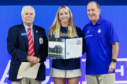 A female student-athlete stands with an acceptance letter from the United States Coast Guard Academy. She wears a blue IMG academy shirt, the man on the left is in a navy blazer with a red striped tie, while the man on the right is in a blue IMG Academy polo shirt.