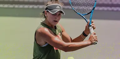 A female tennis player prepares to hit a backhand shot, focusing on the incoming ball while holding her racket with both hands on an outdoor court.