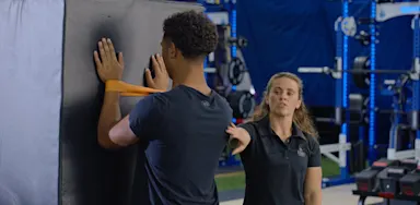 An athlete presses his hands against a wall with a resistance band around his wrists as a trainer observes.