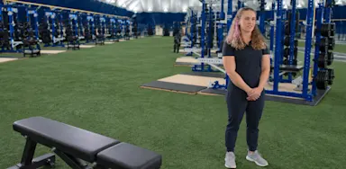 A trainer stands on indoor turf beside a bench and weight racks.