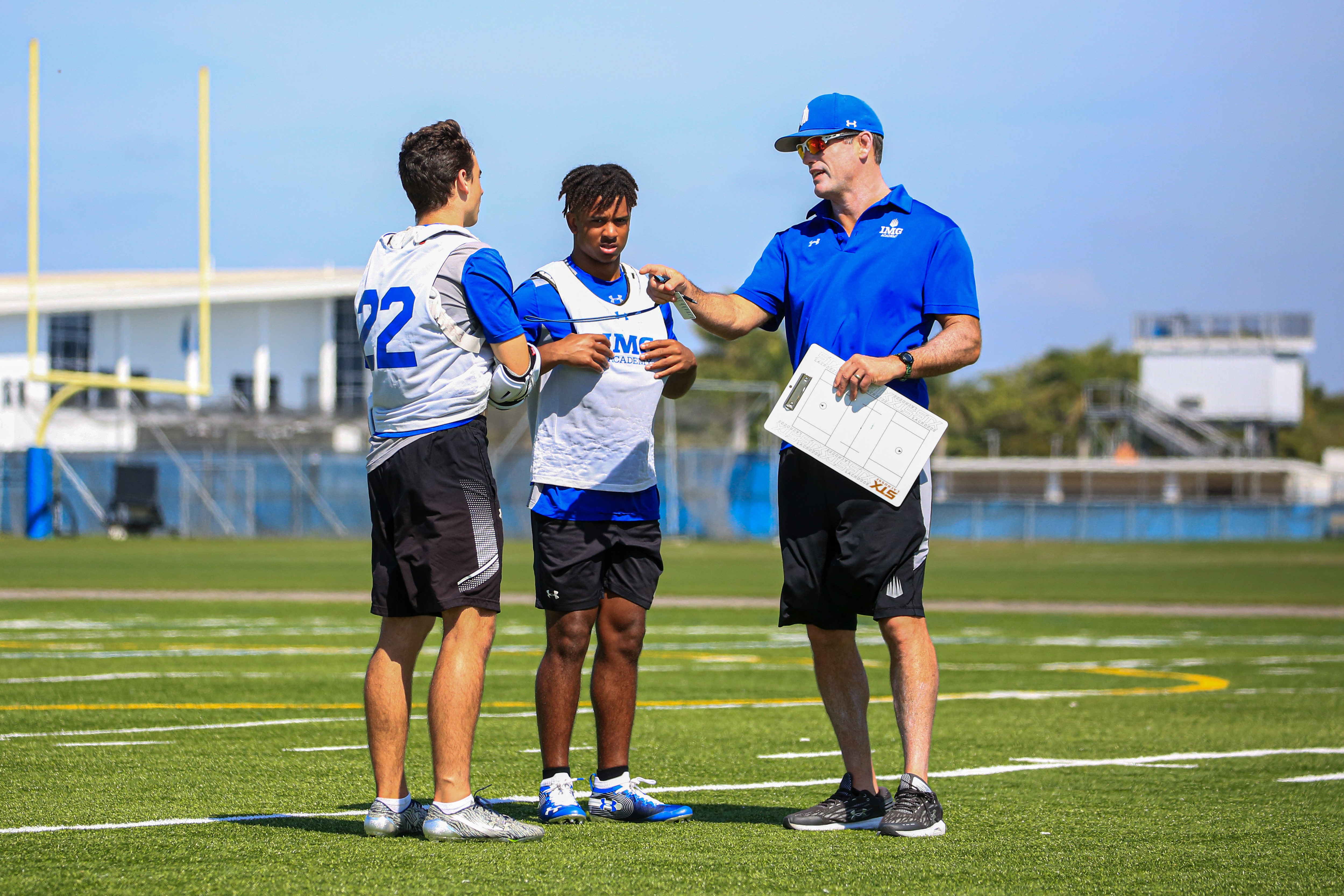 Coach speaking with two student-athletes on a football field during practice, illustrating the importance of communication in the college recruiting process.