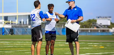 Coach speaking with two student-athletes on a football field during practice, illustrating the importance of communication in the college recruiting process.